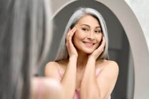 woman with gray hair looking at mirror touching face