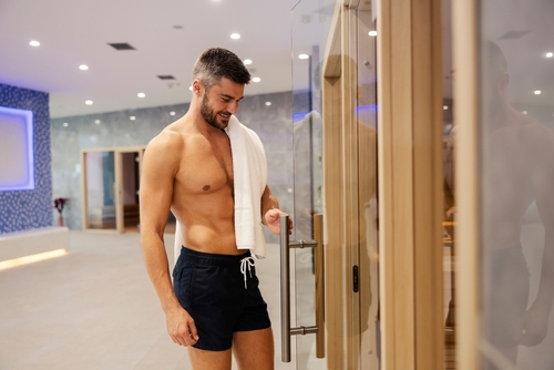 shirtless man in a swimsuit with a towel on his shoulder entering sauna