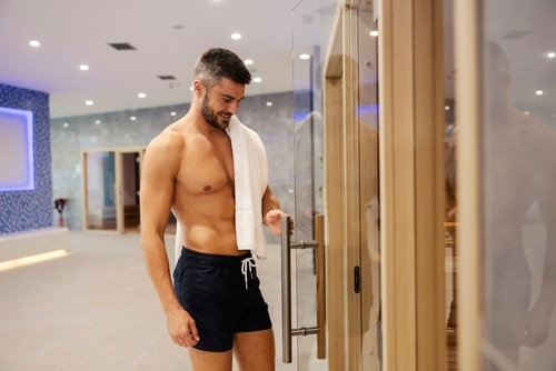 shirtless man in a swimsuit with a towel on his shoulder entering sauna