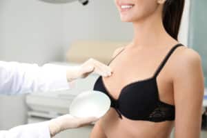Smiling brunette woman discusses breast lift and implants with doctor, holding a silicone implant in his hand